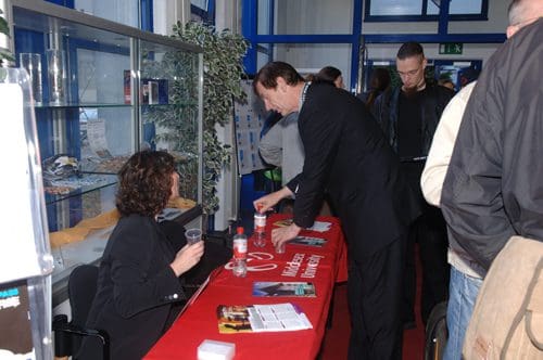 A man in a suit interacts with a woman at a registration table covered with a red cloth, displaying brochures. People are gathered in the background, with display cases of items visible.