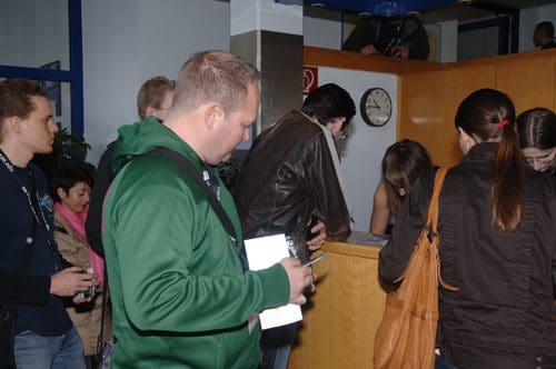 A group of people gathered around a reception desk in a busy lobby, with one person checking in while others are waiting. The scene includes a mix of casual and business attire, plants in the background, and a clock mounted on the wall.