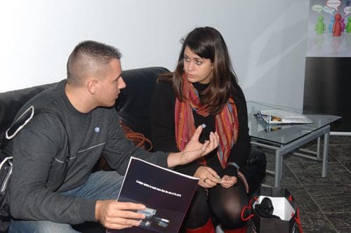 A man and a woman sitting on a black leather couch engaged in conversation. The man, wearing a gray sweater, holds a dark booklet, while the woman, dressed in a black top and colorful scarf, listens attentively.