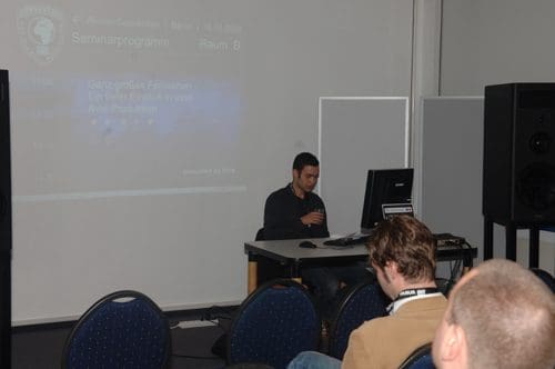 A speaker is sitting at a desk with a computer in a seminar room, preparing for a presentation. The audience, seated in blue chairs, is facing the speaker, while a screen displays the seminar program in the background.