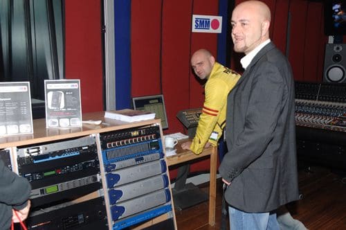 Two men in a music studio. One man in a black blazer smiles and stands near a table with audio equipment. The other man, wearing a yellow jacket, adjusts a device at a workstation in the background, surrounded by studio equipment and sound panels.