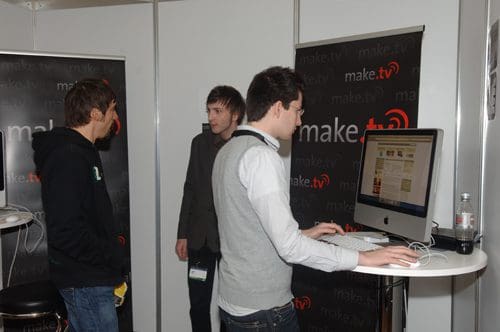 Three individuals at a technology exhibition booth, engaged in conversation near a large computer displaying a website. One person stands with a banana, while another examines the screen attentively.