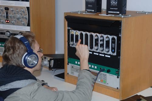 A person wearing headphones operates a sound mixing console, adjusting various knobs and buttons on the front panel. The setting includes audio equipment in the background, emphasizing a sound technology environment.