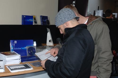 Two individuals examining material at a display. One person in a gray hat holds a booklet while standing at a table cluttered with brochures, CDs, and publications, showcasing music software and resources.