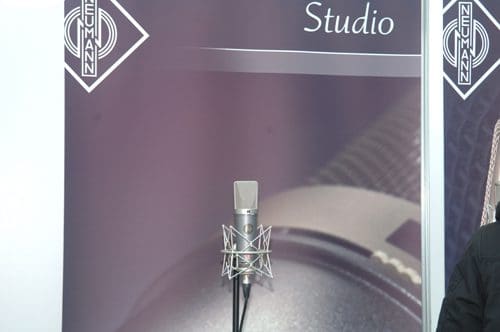 A professional studio microphone mounted on a stand, positioned in front of a backdrop featuring the Neumann logo, with soft gradients in shades of purple and gray.
