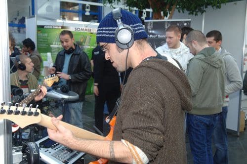 A man playing an electric guitar while wearing headphones at a music exhibition. Several people are gathered around, listening and observing the demonstration, with various music equipment displayed in the background.