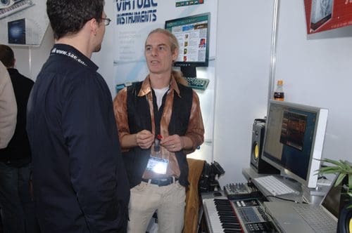 Two men engaged in conversation at a technology exhibition. One man, wearing a black shirt, faces another man with long hair, dressed in a patterned shirt and black vest. Various music equipment, including a keyboard and computer, are visible in the background.