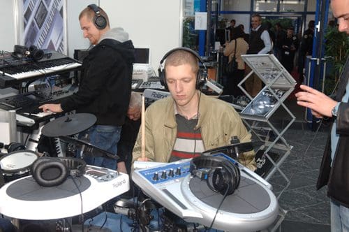 A man wearing headphones plays an electronic drum set with a digital interface while another person adjusts synthesizers in the background. The setting appears to be a music technology exhibition.