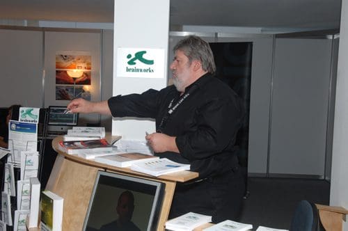 A man with gray hair wearing a black shirt organizes brochures at a wooden reception desk in an exhibition space. Displays and screens are visible in the background, suggesting a professional event.