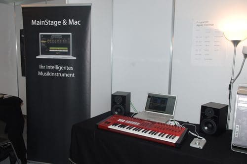 A booth displaying music equipment featuring a large banner with text about MainStage & Mac. On the table, a red keyboard controller is placed alongside a laptop and two studio speakers, all set against a neutral background.