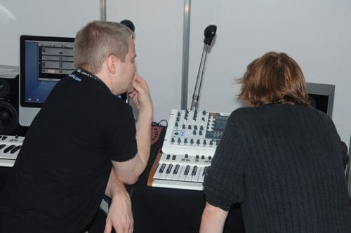 Two individuals intently focused on a music production setup. One man, wearing a black shirt, rests his chin on his hand while examining a synthesizer. The other person, with shoulder-length hair, observes the equipment from the side.