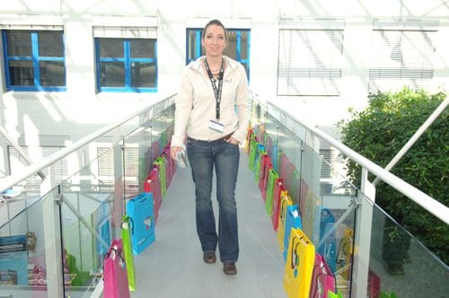 A woman walks along a glass bridge lined with colorful bags hanging on each side, surrounded by bright natural light from large windows. Lush greenery is visible in the background, enhancing the vibrant atmosphere.