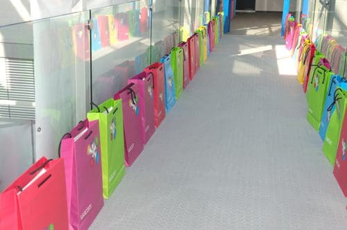 A narrow hallway lined with colorful gift bags in pink, green, and blue, arranged neatly along the floor. The walls are bright, enhancing the cheerful atmosphere of the space.