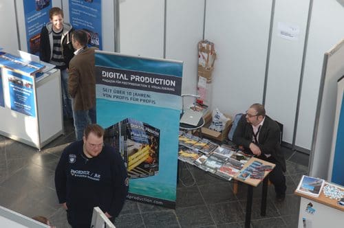 A trade show scene featuring a booth for 'Digital Production' with a large banner. Two men are engaged in conversation at the booth, while another man stands nearby, looking at printed materials on a table.