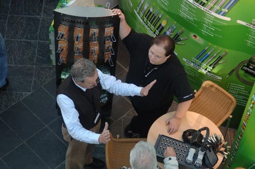 Two men engage in a discussion at a trade show booth, surrounded by green promotional materials and products. One man gestures with his hands while explaining, and the other listens attentively, seated beside a table with various items.