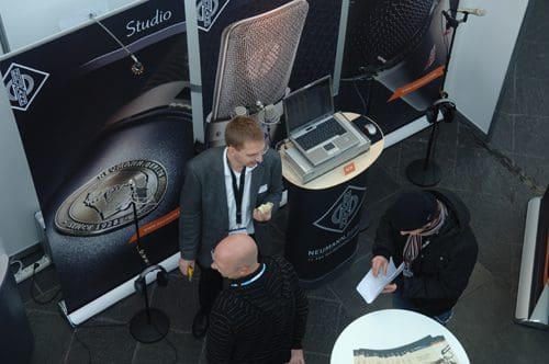 An overhead view of a tradeshow booth featuring a man in a gray blazer speaking with another person. Background displays showcase microphone products. A laptop sits on a table next to promotional materials.