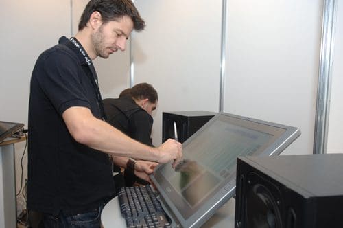 A man using a large touchscreen display, drawing with a stylus while standing beside a keyboard. The background shows another person focused on their tasks, with audio speakers positioned nearby in a modern workspace.