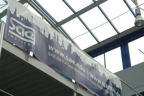 A large banner hanging from a ceiling, displaying the SAE Institute logo and website addresses. The background features a silhouette of a city skyline. The overall atmosphere is bright and open, with natural light filtering through the glass roof.