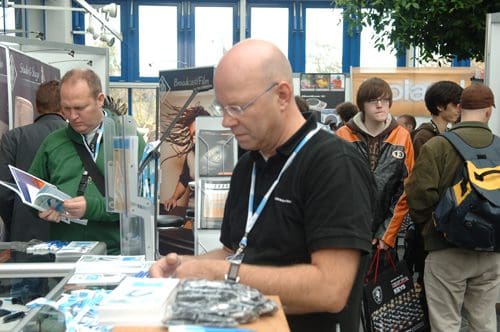 A man wearing glasses browses through promotional materials at a trade show booth, while other attendees engage in conversation and explore nearby displays in a busy exhibition space.
