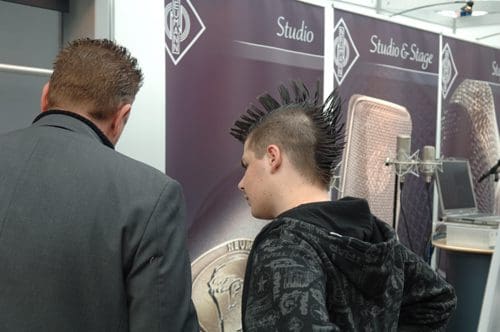 Two individuals are standing with their backs to the camera, discussing something. One has a spiked mohawk hairstyle, and is wearing a dark hoodie. The other, dressed in a gray suit, appears engaged in the conversation. A backdrop features branding related to studio equipment.