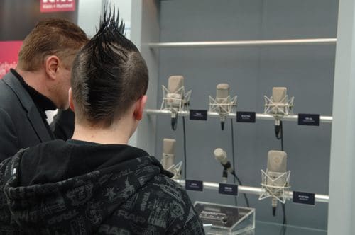 Two individuals viewing a display of microphones in a technology exhibition. One has a mohawk hairstyle while the other wears a suit, both focused on the various microphone models arranged on a shelf.
