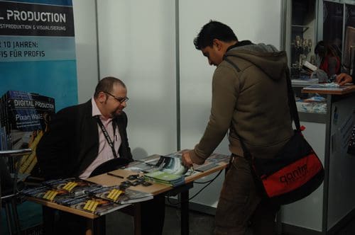 A man in a dark jacket interacts with a visitor at a trade show booth. The booth showcases promotional materials, with a sign visible in the background, indicating a focus on digital production.