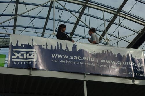 Two individuals converse on a balcony, overlooking a banner featuring the silhouette of a city skyline and promoting the SAE Institute. The roof structure has a glass architecture.
