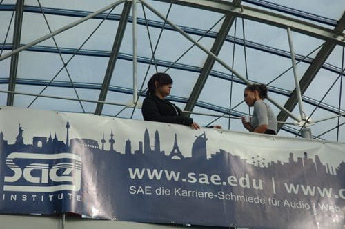 Two individuals engaged in conversation on a balcony, surrounded by a large banner featuring landmarks in silhouette, beneath a glass roof with metal supports.