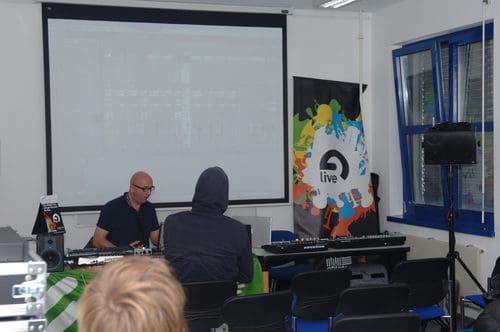 A music workshop setting with a man sitting in front of a computer screen, demonstrating music production software. Two attendees, one in a hoodie, watch from their seats. A colorful banner featuring the Ableton Live logo is displayed on the side.