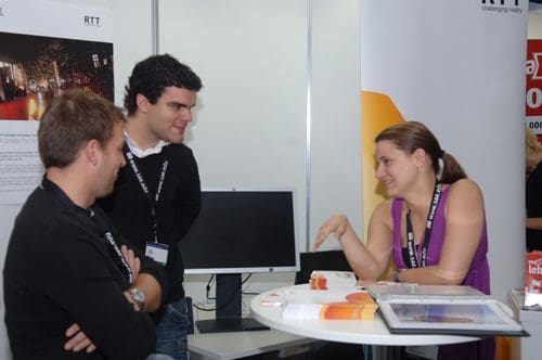 Three individuals engaged in conversation at a professional exhibition booth. A woman gestures while explaining, while two men listen attentively. A computer and promotional materials are visible on the table.