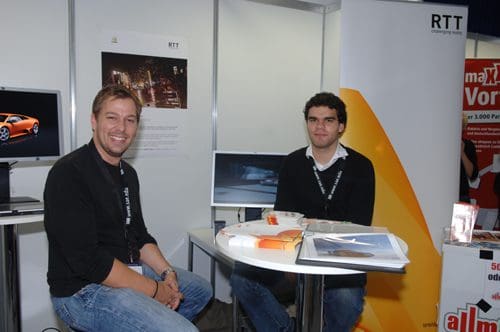 Two men seated at a table in a trade show booth. One man, on the left, smiles while wearing a black shirt and jeans. The other man, on the right, is dressed in a black sweater and is engaged in conversation. Behind them are monitors displaying images.