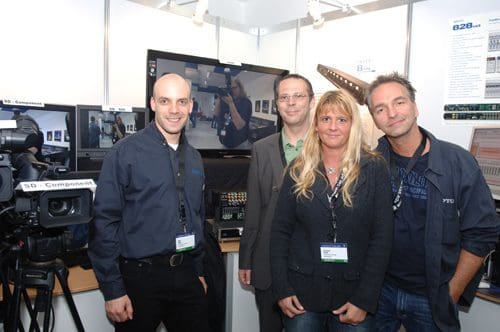 Four individuals pose for a photo at a technology exhibition booth, with a camera and equipment. They are smiling, dressed in semi-formal attire, and standing in front of a large screen displaying technical content.