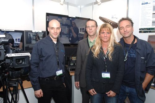 Four professionals posing at a trade show booth, with video equipment and monitors in the background. One man wears a dark shirt, another in a suit, a woman in a black jacket with long hair, and a man in a casual shirt. They all smile at the camera.