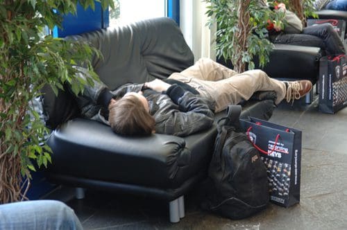 A young person reclining on a black leather couch, with legs stretched out and a backpack resting beside them, in a casual indoor waiting area adorned with potted plants.