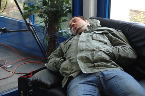 A man wearing a green jacket and jeans is sleeping on a black leather couch in a well-lit room. He has headphones on and his hands are tucked into his jacket pockets, with a plant visible in the background.