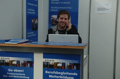 A young man sitting at a table with a laptop, engaged in a phone call at a trade fair booth. The booth displays colorful brochures and a banner promoting professional training and development opportunities.