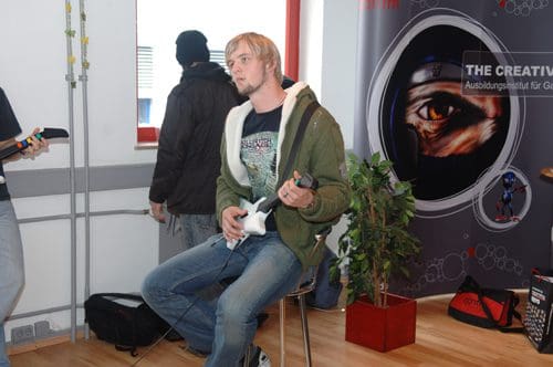 A young man sitting on a stool, playing a guitar video game with a controller, in a modern room. Another person in a black jacket stands in the background near a window. A potted plant is visible beside them, and a promotional banner is on the wall.