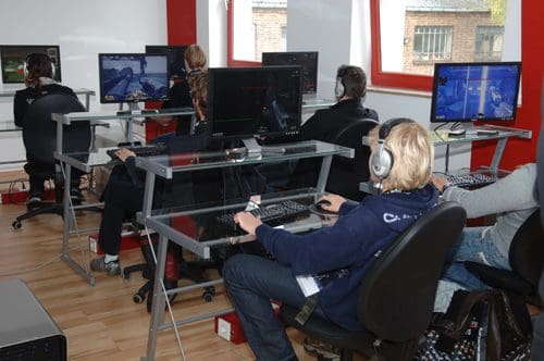 A group of people seated at modern glass desks, wearing headphones while focused on computer monitors displaying video games. The workspace has a bright, contemporary look with large windows and wooden flooring.