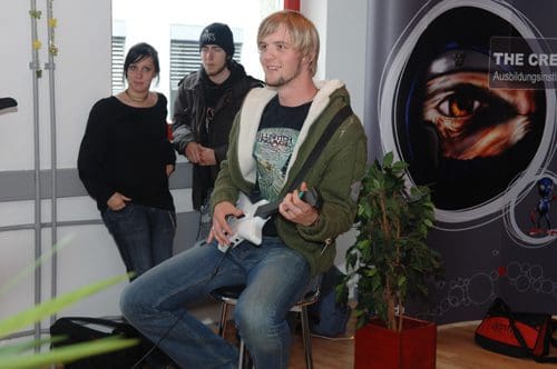 A young man sitting on a chair, holding a guitar controller, smiling while playing a video game. Two spectators watch him, one woman in black attire and a man in a beanie and jacket, with a colorful backdrop featuring a design.