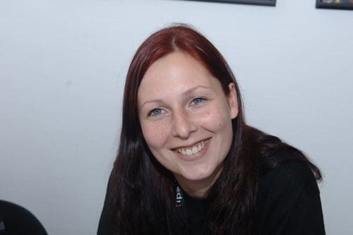 A woman with long, dark hair and a cheerful smile sits at a table, wearing a black t-shirt. The background consists of a plain white wall and framed pictures, creating a friendly and inviting atmosphere.