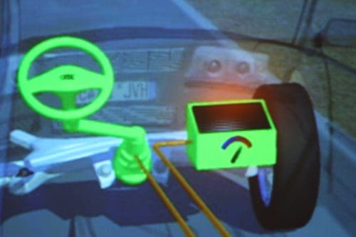 A digital display showing the steering mechanism of a vehicle, featuring a bright green steering wheel connected to a tire. An indicator box displays operational parameters with a gauge, all set against a blurred road backdrop.