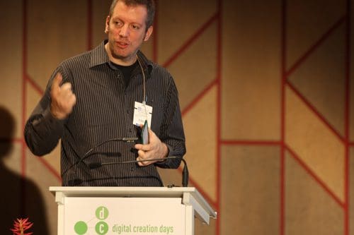 A speaker gesturing with one hand while presenting at a conference. He is wearing a striped black shirt and holding a device in his other hand, with a podium displaying the text 'digital creation days' in the foreground.