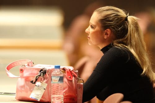 A woman with long hair styled in a ponytail sits at a table, looking thoughtfully away. Beside her, a pink handbag and a water bottle are placed on the table in a softly lit venue.