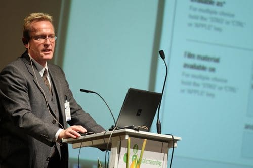 A man in a business suit presenting at a conference, standing behind a podium with a laptop. A projection screen behind him displays text related to film material availability.
