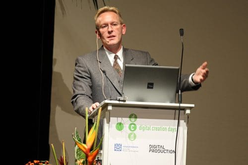 A speaker presenting at a conference, standing beside a podium adorned with flowers. He wears a gray suit and adjusts a microphone while gesturing with his right hand, with a laptop visible on the podium.