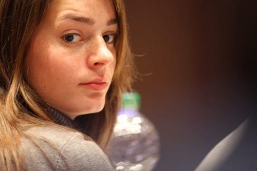 A young woman with long brown hair looks thoughtfully over her shoulder while sitting at a table. A blurry water bottle is in the background, creating a focused and contemplative atmosphere.
