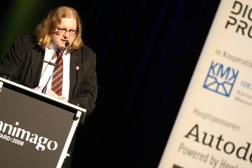 A man with long hair and glasses stands at a podium, delivering a speech during an award ceremony. He wears a suit and a striped tie, with a logo banner featuring sponsors in the background.