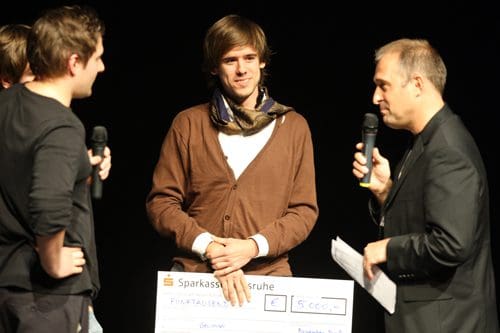 A man wearing a brown sweater and scarf smiles while holding a large check on stage. Two other individuals stand nearby, one speaking into a microphone and another holding a microphone. The backdrop is dark, emphasizing the moment.