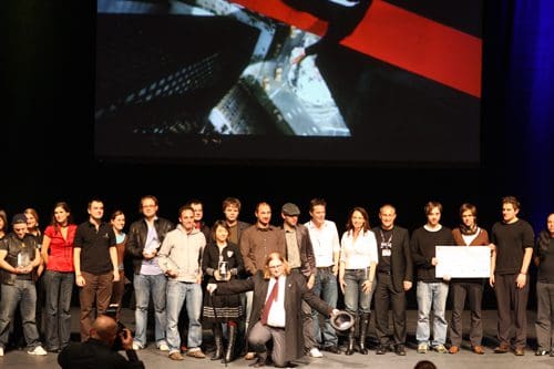 A diverse group of people standing on stage, celebrating an achievement. They are positioned in front of a large screen displaying a colorful visual. Some individuals hold trophies and awards, while others smile and pose for the camera.