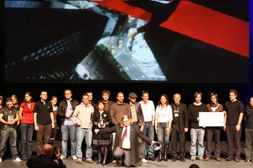 A diverse group of people standing together on stage during an awards ceremony, with a large screen in the background displaying a vibrant visual. Some individuals are holding a large check and trophy.